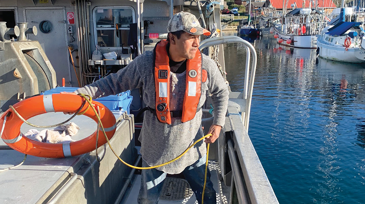 A commercial fishing worker wearing a life jacket stands on the deck of a fishing vessel, handling a rope beside a life ring, with other fishing boats moored in a harbour in the background