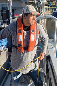 A commercial fishing worker wearing a life jacket stands on the deck of a fishing vessel, handling a rope beside a life ring, with other fishing boats moored in a harbour in the background.