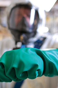 Worker wearing protective gloves and coveralls uses a hand tool in an industrial setting.