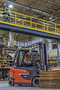A forklift operating inside West Fraser’s Williams Lake Plywood facility while a worker walks along an overhead catwalk, illustrating pedestrian safety around mobile equipment on work sites.
