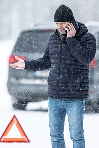 A person wearing a dark toque, dark winter jack, and blue jeans standing beside a car in falling snow, talking on a phone with a roadside warning triangle on the ground.