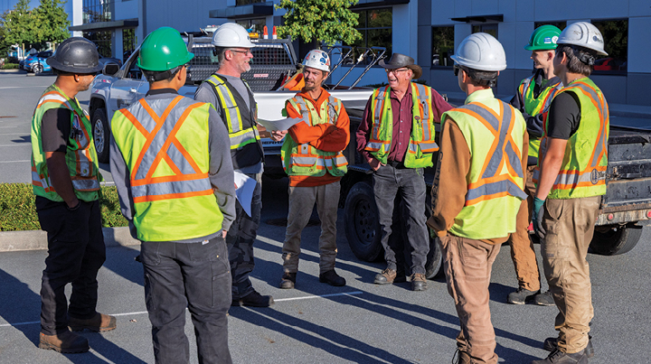 Roadside workers wearing high-visibility vests and hard hats stand in a group during a safety talk.