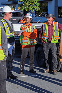 Roadside workers wearing high-visibility vests and hard hats stand in a group during a safety talk.