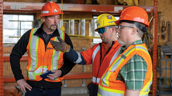 Three workers wearing hard hats and high‑visibility safety vests standing inside an industrial workshop, with one worker gesturing while speaking to the others, surrounded by shelving, tools, and construction materials.
