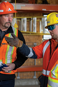 Close up of two workers in hard hats and safety vests having a conversation. The worker on the right is gesturing forward with a gloved hand.