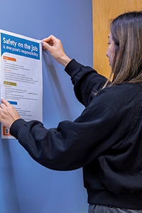 Close up of a person hanging a poster on a wall. The text on the poster reads "Safety on the job".