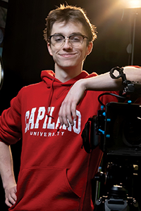 Image of a young person wearing glasses and a red sweatshirt. Their elbow is raised and leaning on camera equipment.