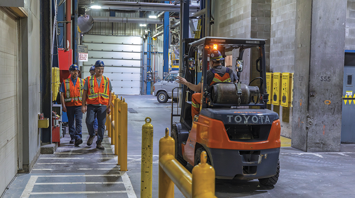 A forklift operating in an industrial facility as workers in high-visibility gear walk nearby along a designated pedestrian pathway, illustrating measures to prevent of struck-by incidents around mobile equipment.