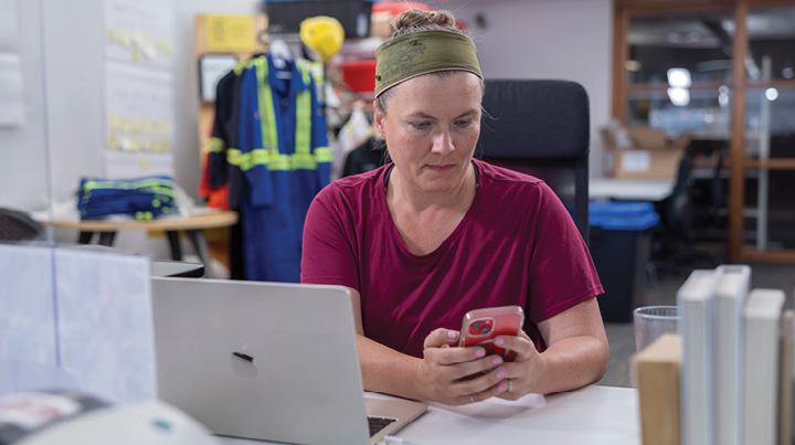 Worker seated at a desk using a mobile phone beside an open laptop in an office setting.