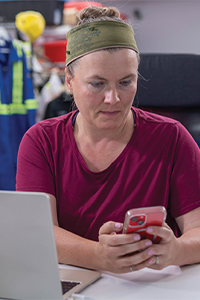 Worker seated at a desk using a mobile phone beside an open laptop in an office setting.