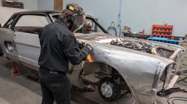 Worker wearing protective equipment grinding a metal vehicle panel in an automotive workshop.