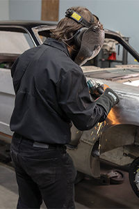 Worker wearing protective equipment grinding a metal vehicle panel in an automotive workshop.