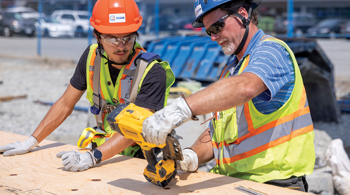 Two construction workers wearing hard hats and safety vests using a power tool on a plywood work surface.