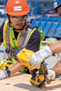 Two construction workers wearing hard hats and safety vests using a power tool on a plywood work surface.