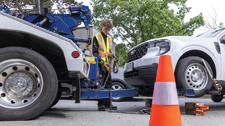 Worker in high-visibility safety gear securing a vehicle onto a tow truck bed with traffic cones in the foreground.