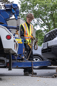 Worker in high-visibility safety gear securing a vehicle onto a tow truck bed with traffic cones in the foreground.