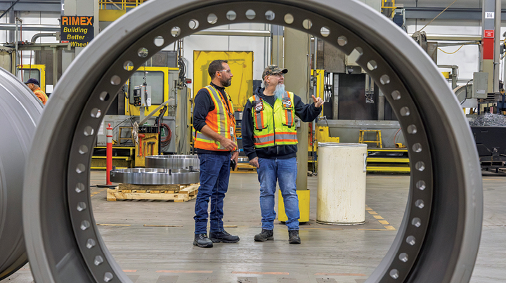 Two workers wearing high‑visibility safety vests standing inside a manufacturing facility, viewed through a large circular metal component in the foreground, with industrial equipment and machinery surrounding them.