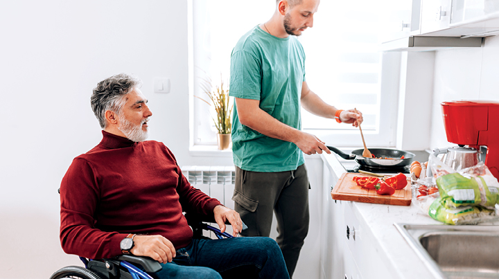 Two people in a bright kitchen, with one person cooking food in a frying pan on a stove while another person seated in a wheelchair observes, with vegetables and kitchen appliances visible on the counter.