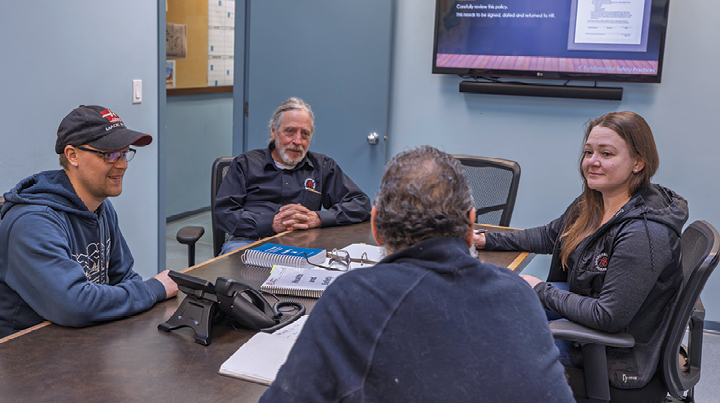 Four people seated around a meeting table in an office, reviewing documents and discussing materials, with a wall-mounted screen and office supplies visible in the room.