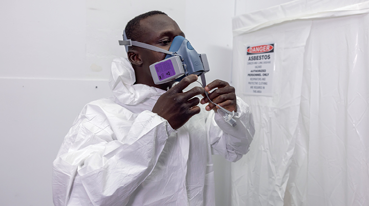Worker wearing full protective coveralls and a respirator adjusting the mask inside a controlled environment area, with a posted warning sign indicating asbestos hazard on a sealed doorway
