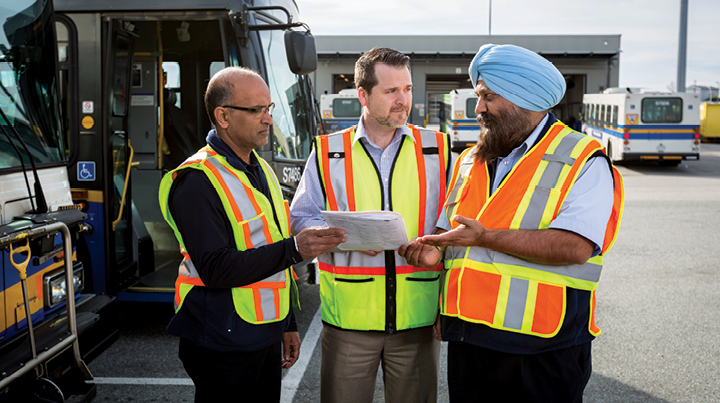 Three workers wearing high‑visibility safety vests standing beside a transit bus, reviewing a document together in an outdoor vehicle yard with additional buses in the background.