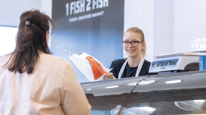 Retail worker behind a counter handing a piece of paper-wrapped food product to a customer across a glass display case, with signage reading ‘1 FISH 2 FISH’ visible on the wall behind.