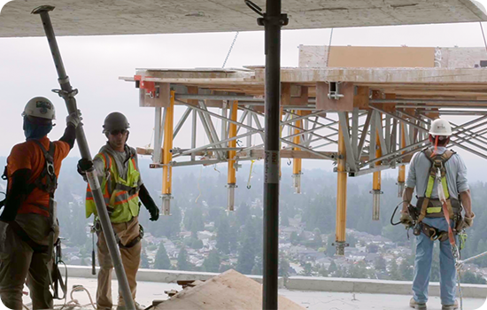 Construction workers wearing fall protection, hard hats, and high-vis vests watch a flytable approaching the floor they're standing on